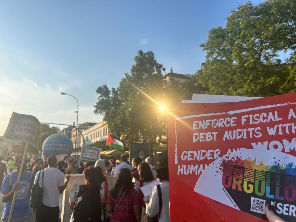 Image with people protesting with a "Cancela la deuda" (Cancel the debt) sign and a Palestinian flag. 