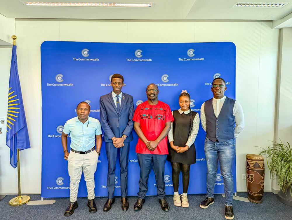 A group of 5 people in casual and formal attire posing in front of a Commonwealth step and repeat banner smiling to the camera.
