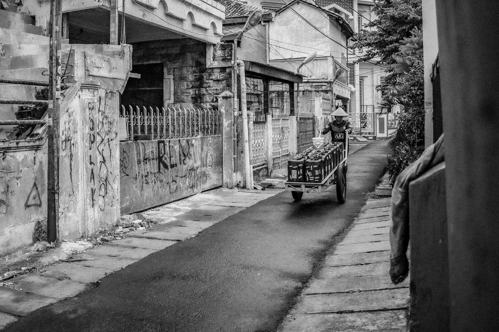 A black and white photo by D. Arifah, titled &quot;Water Bearers&quot;, in which a man in a pointed sun hat pushes a handcart loaded with plastic water containers down an alley lined with fences and graffiti-covered walls.