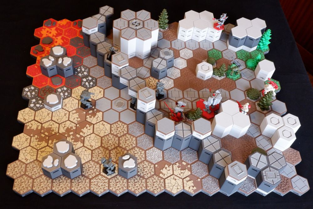 A table of 3D printed hex terrain, depicting a city area protected by a wall, outside which is dry ground and an encroaching lava flow.  Mech miniatures face each other through the gate in the wall.