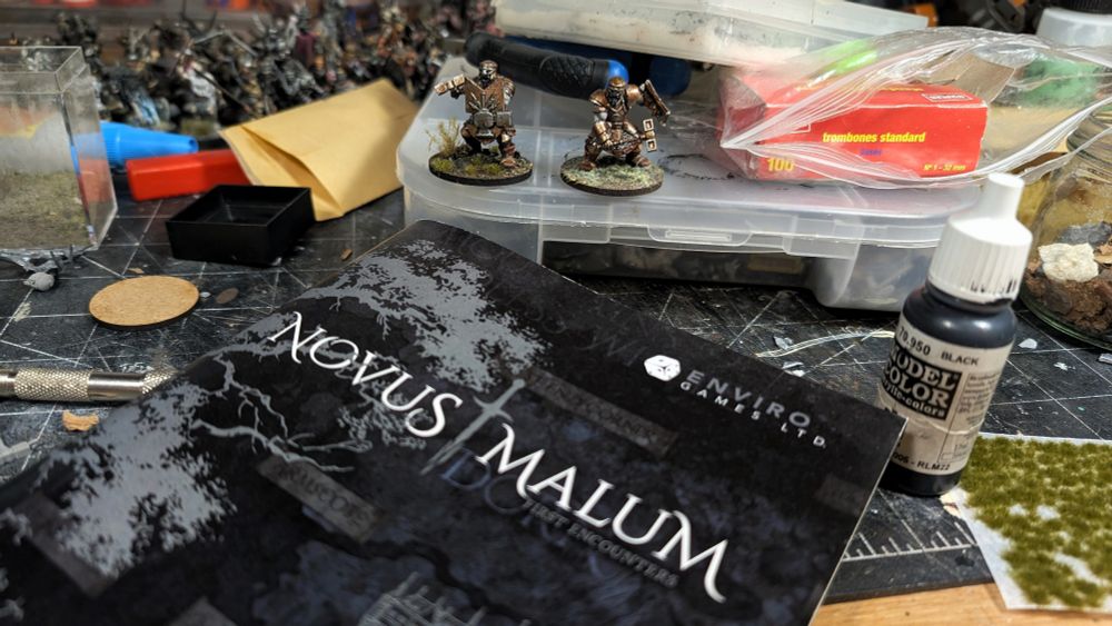A couple painted models sitting on a pile of stuff on a messy book with a small dark grey book lies in front.  Books is titled Norvus Malum by Enviro Games ltd