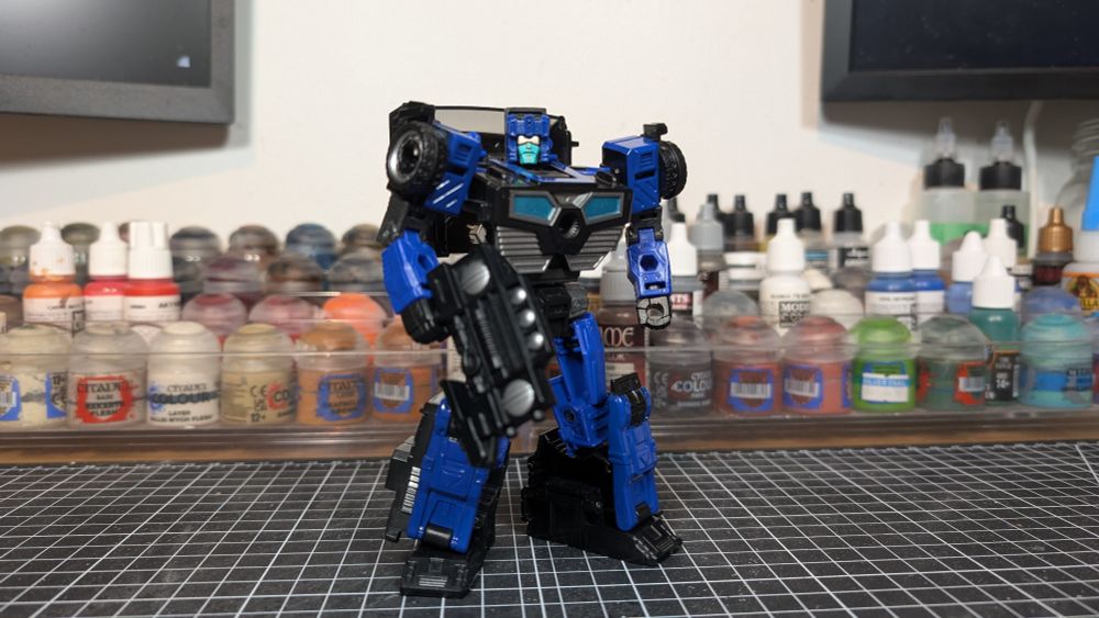 Blue and black robot toy on painting desk with cutting mat, and paint pots on racks behind him