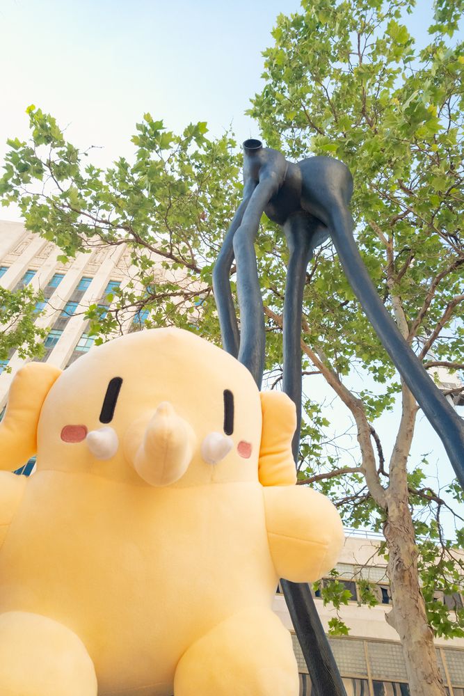 Photo of the Creature Mastodon stuffed toy standing beneath a towering, unusual quadruped sculpture outdoors in Los Angeles during the day. The Creature is in the foreground, facing down at the camera, which is pointed upwards toward one of the monkeys among the “sixbeastsandtwomonkeys” sculptures by Peter Shelton.