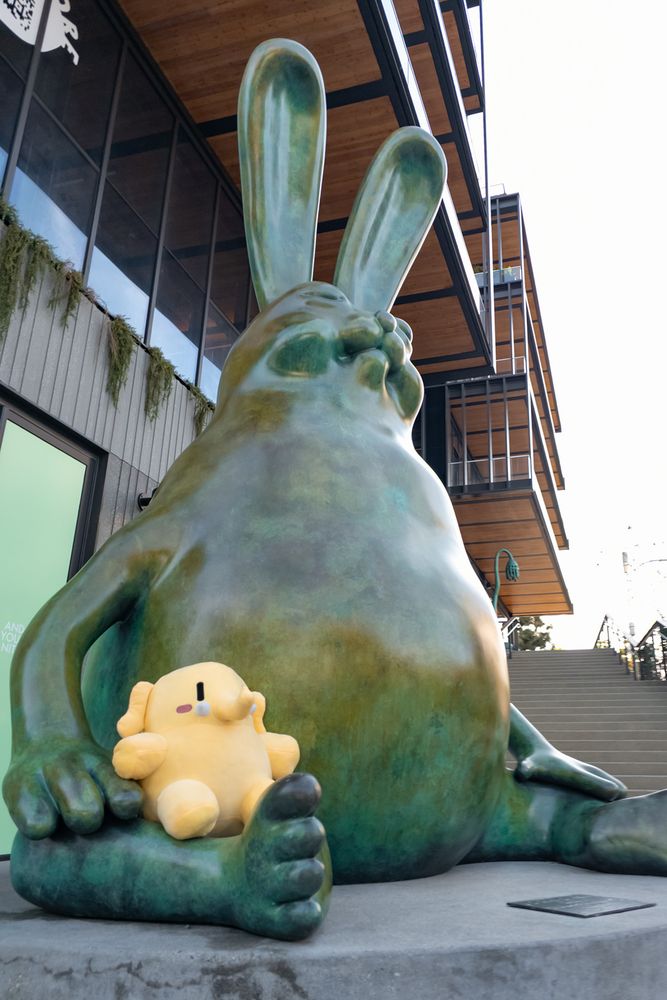 Photo of the Creature Mastodon stuffed toy sitting on a giant, unusual rabbit sculpture outdoors in Los Angeles during the day. The Creature's sitting on the lap of the Be Hare Now sculpture by the Haas Brothers, which is sitting on a concrete platform, as they both stare up towards image top-right.