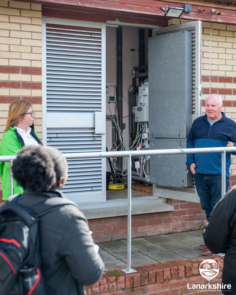 Two people standing outside an electrical cupboard speaking with other people in a crowd.