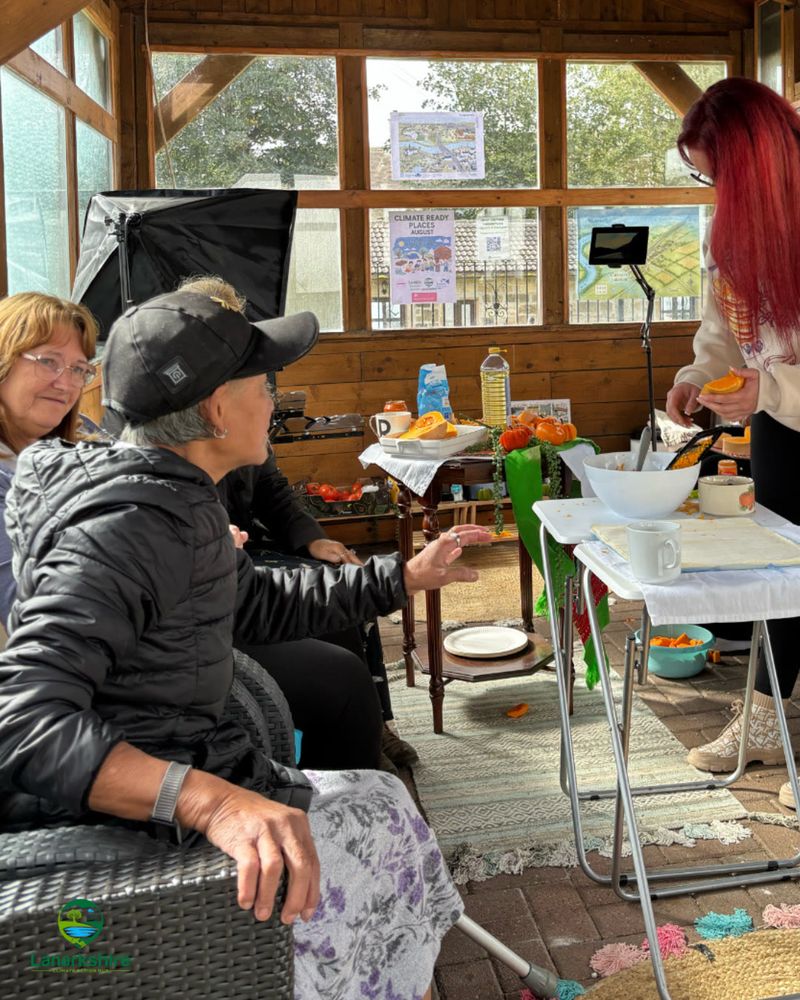 One person preparing food at a table with three people sitting on a sofa watching.
