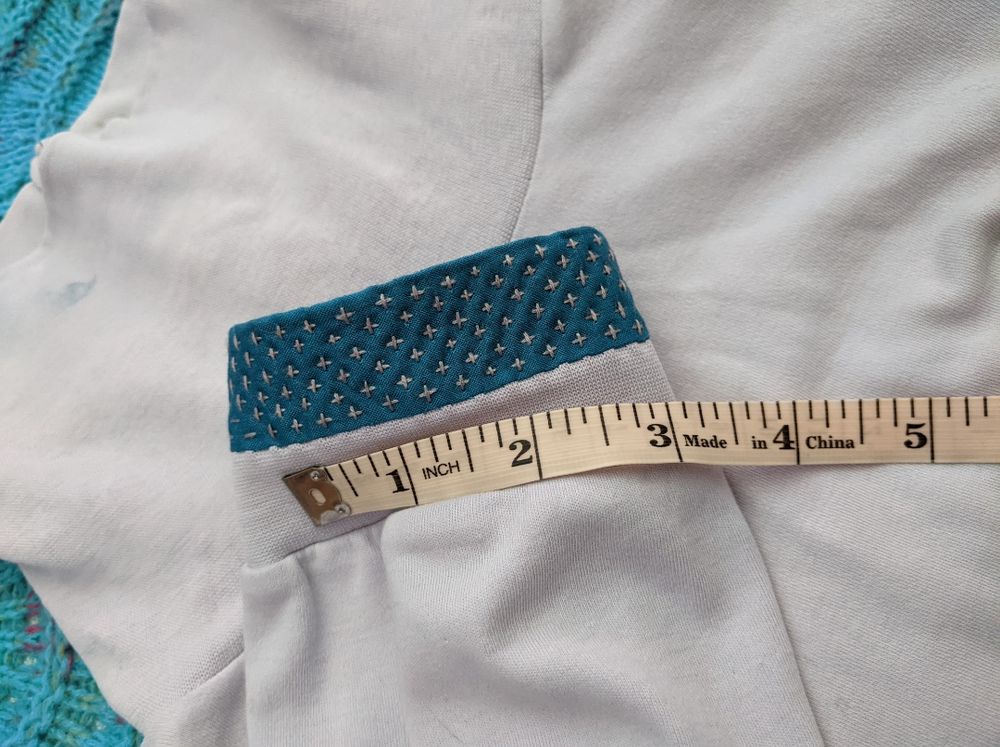 Picture of a light lavender turtle neck with dark teal fabric sewn onto a sleeve cuff as visible mending. A tape measure shows how small the light purple stitches are: around 4 every inch.