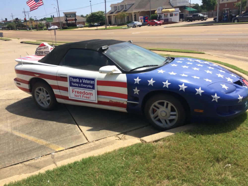 A US flag wrapped convertible Pizza Hut delivery car with a Thank A Veteran Freedom Isn’t Free sign on the passenger door. 