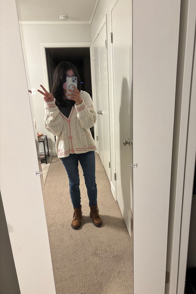 Mirror selfie dressed in gray shirt, dark blue jeans, brown ankle boots, and Taylor Swift Lover cardigan (white knit cardigan with light pink trim, embroidered hearts, and three light pink heart buttons)
