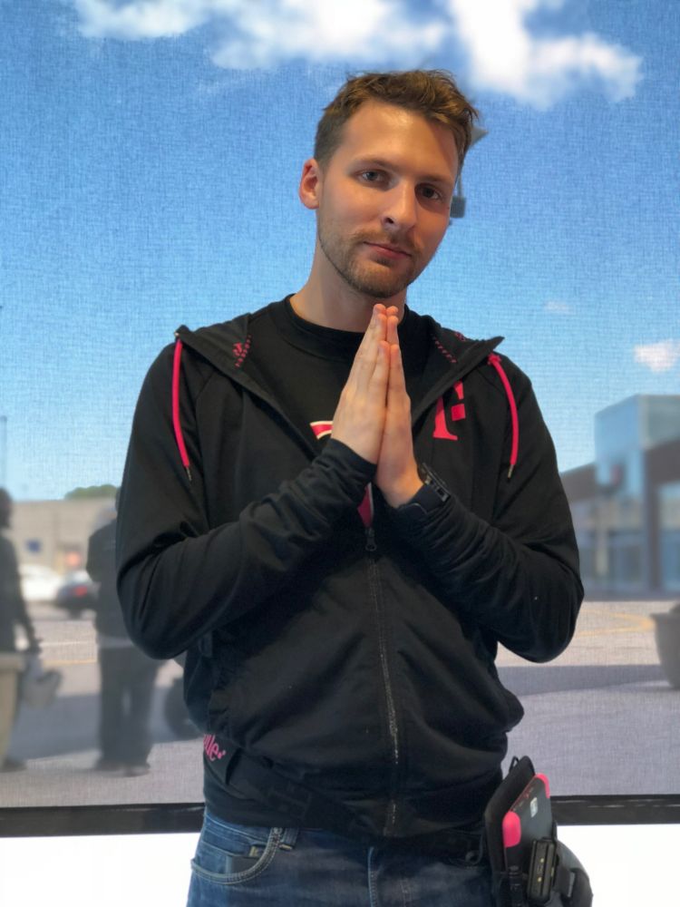 pre-transition photo of lyra in t-mobile retail uniform circa 2017 with short hair and visible facial hair (scruff?) with both hands in a sort of praying pose