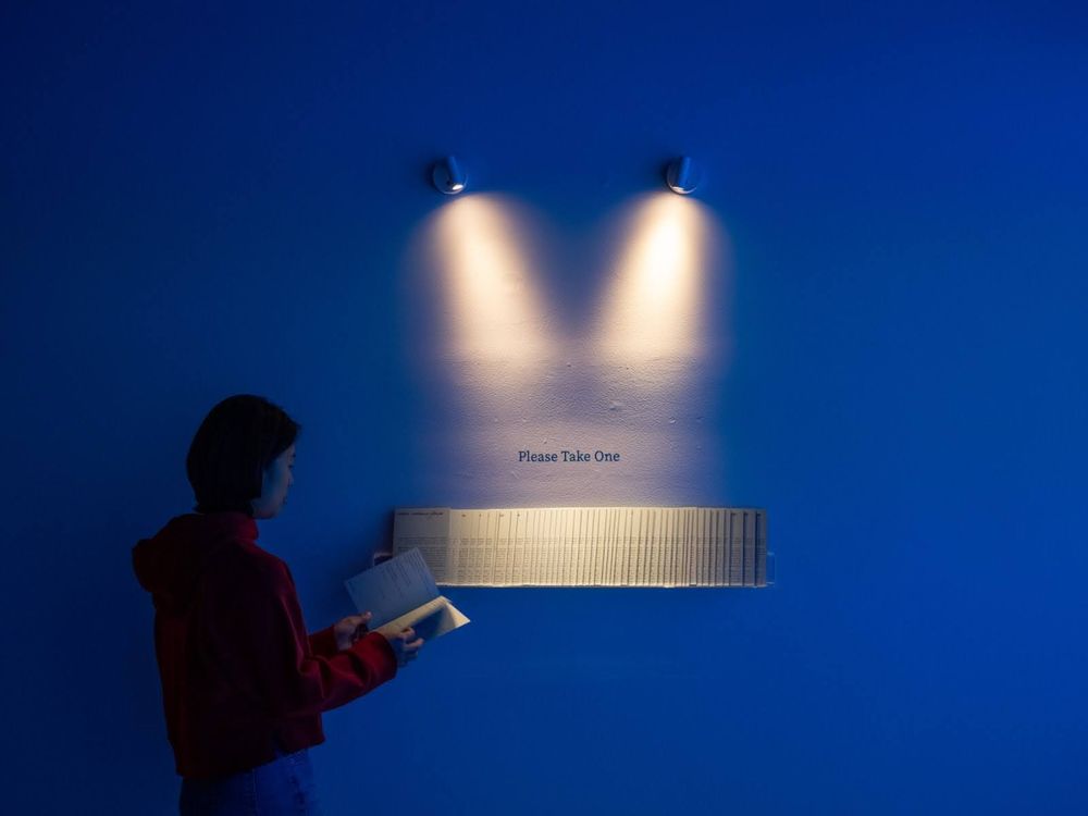 Installation View of where violence flows (2025), with a row of zines under two spotlights along with a sign saying, "please take one." Photo by Andy Kajie.