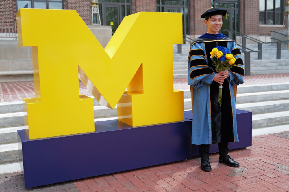 Man in doctorate regalia and holding a bouquet of flowers next to a giant block M in front of the Hatcher Library.