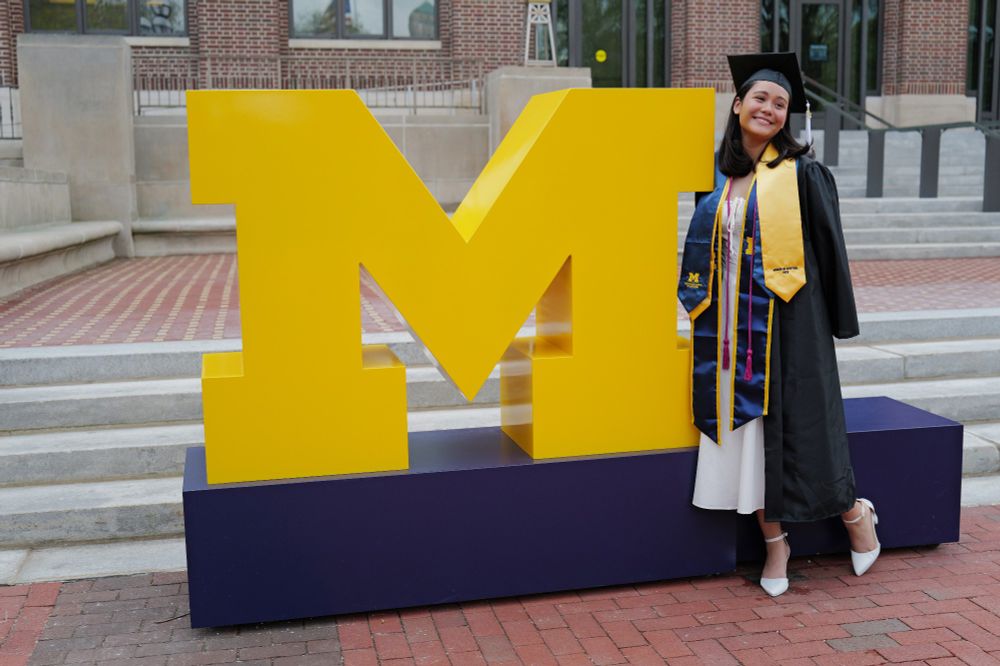 Woman in graduation gown and stoles posing next to a giant block M in front of the Hatcher Library.