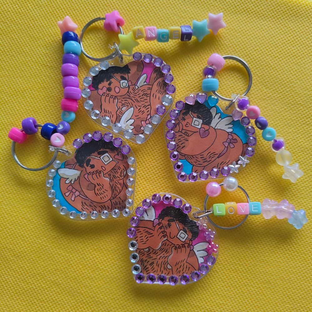 Photo of four photo holder keychains with drawings of my original character Allan posing. The holders are decorated with sparkling stickers and beans.