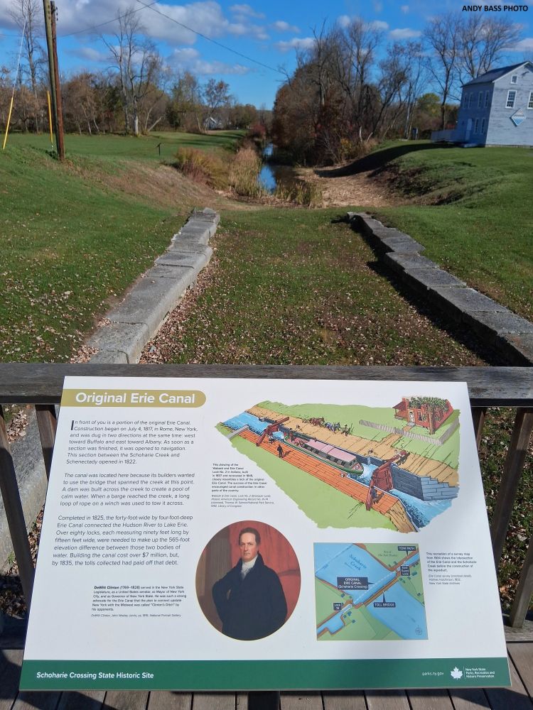 An observation deck with a wayside exhibit titled, “Original Erie Canal,” overlooks a surviving half-mile-long canal prism of the original Erie Canal at Schoharie Crossing State Historic Site in Fort Hunter, New York.  The stone remains of the East Guard Lock are visible in the foreground.  The original 40-foot-wide canal, known as “Clinton’s Ditch,” was in operation here from 1822 to 1845.  The flotilla led by Governor DeWitt Clinton’s boat Seneca Chief passed through this canal prism on November 1, 1825.  (Andy Bass photo, 2025)