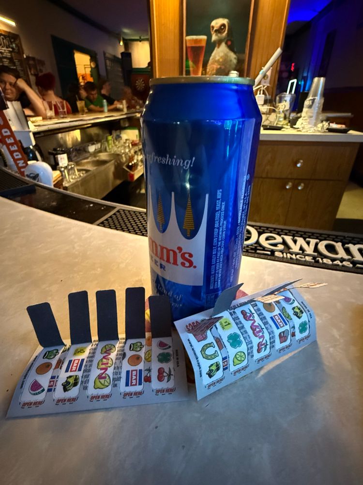 Two winning pull tabs lay at the base of a Hamms Tall Boy. A portrait of a stoic baby falcon drinking a beer is in the background