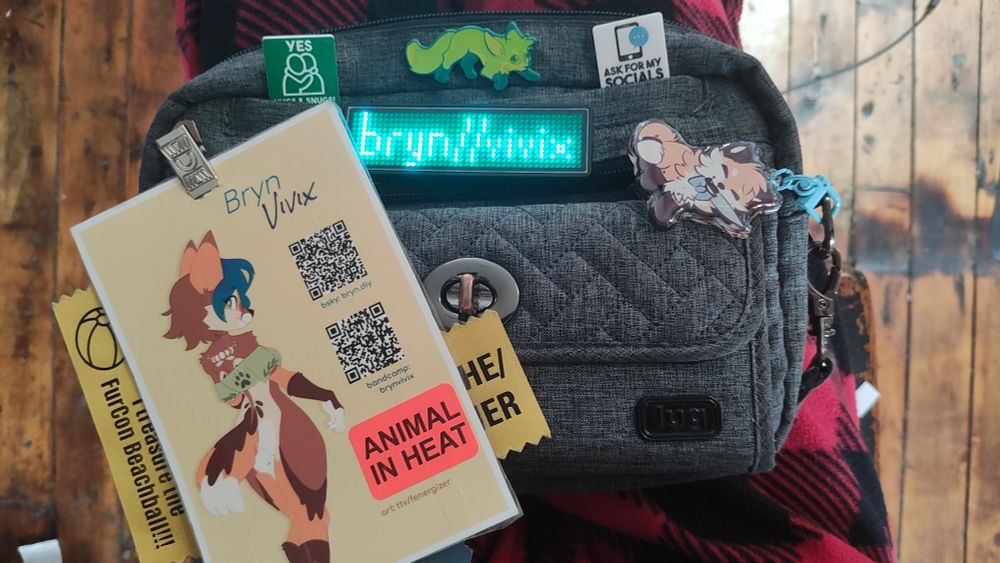 The front of Bryn's blue-grey handbag, adorned with various pins and trinkets. These include:

- Bryn's custom name badge, consisting of her fursona (by @foxbeans.bsky.social), QR codes for her Bluesky and Bandcamp accounts, a bright orange sticker that says "ANIMAL IN HEAT," and ribbons that read "SHE/HER" and "I treasure the FurCon Beachball!!! [sic]"
- a teal LED nametag which currently reads "bryn//vivix"
- Two consent pins: one in green with the 🫂 emoji printed on it and the text "YES HUGS & SNUGS!" and one in white with a cell phone/speech bubble that reads "ASK FOR MY SOCIALS"
- an enamel pin of a red fox, palette swapped to a lime green, slinking forward cautiously. This one is by Ar.Gi.Bi Creative Studio.
- a keychain of a small red fox wearing a blue bowtie and holding a butcher's knife in its mouth in an adorably threatening manner. This one comes from Trashmutt (Kota).
- 