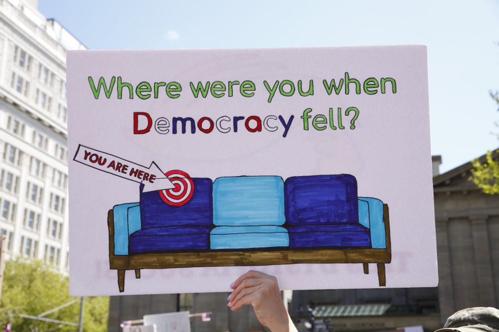 A political sign being held up in Portland, OR's Pioneer Courthouse Square with a drawing of a couch with blue cushions stating, "Where were you when Democracy fell?" With an arrow pointing to a seat on the couch with a target saying, "you are here"