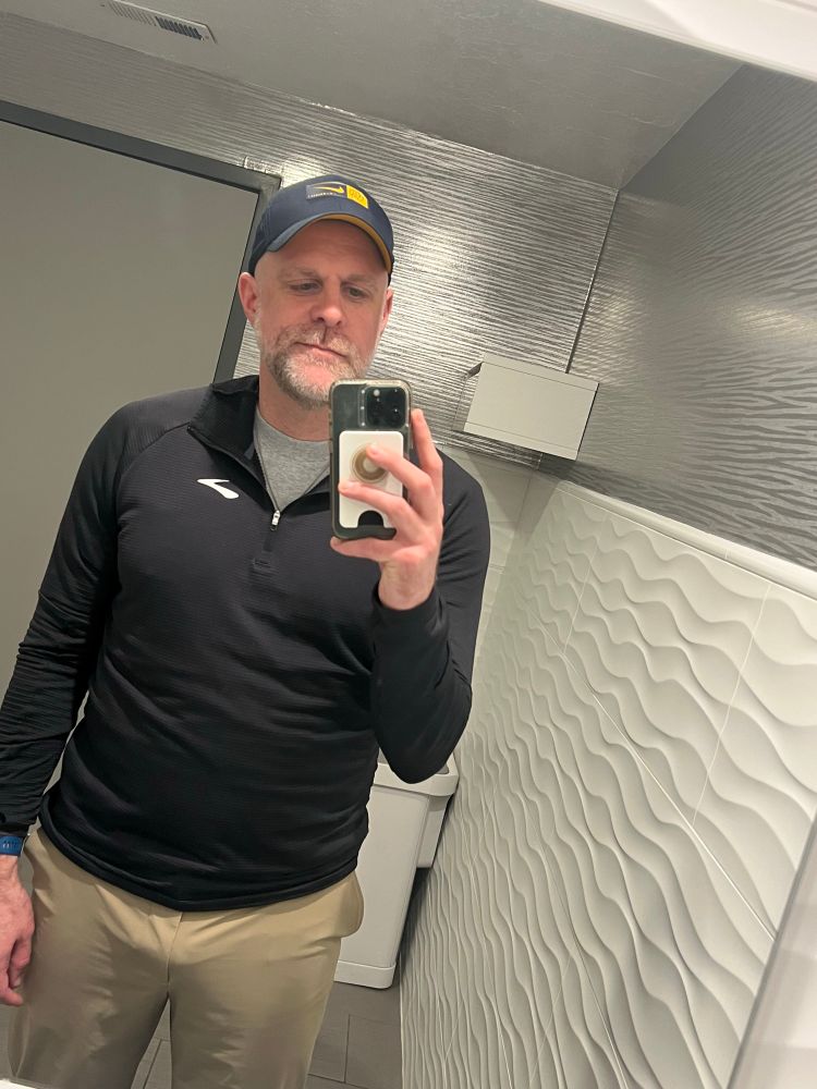 Andrei is wearing an outfit of athleisure wear: a blue Brewers hat, a black Brooks hoodie, and Uniqlo chinos