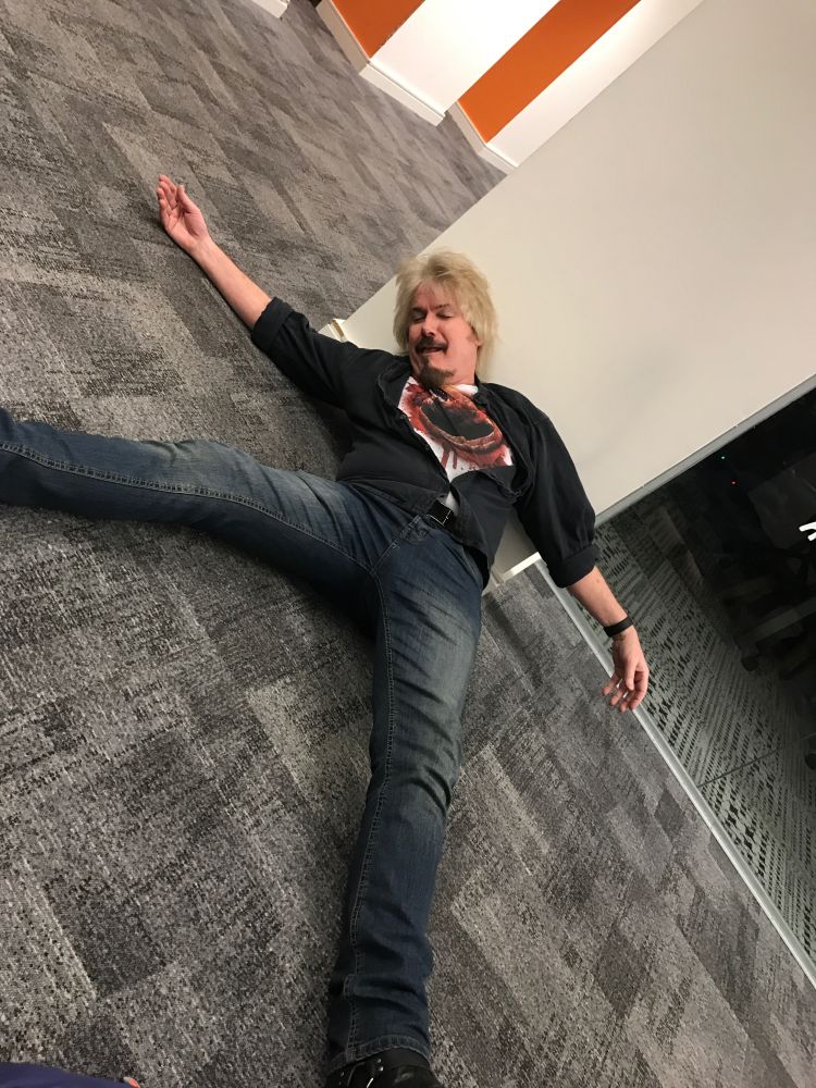 Dirk with eyes closed, spreadeagled on an office corridor floor wearing a tee shirt that makes it look like he's just given birth to an Alien Chestburster.