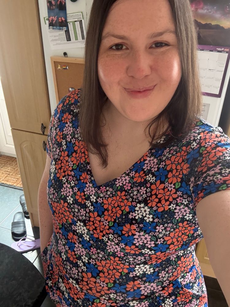 A selfie of me - I have shoulder length brown hair, brown eyes and freckles, I’m smiling and I’m wearing a jumpsuit with flower pattern in red, pink, blue and white colours. 