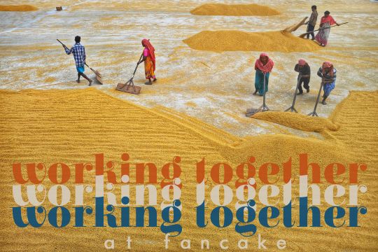 Photograph with added text: Working Together, at Fancake. Workers in India use wide wooden paddles with long handles to shove a huge yard of drying grains into big piles. The grain, most likely rice, is a beautiful golden color, and there's a mix of western and traditional clothing among the seven men and women.