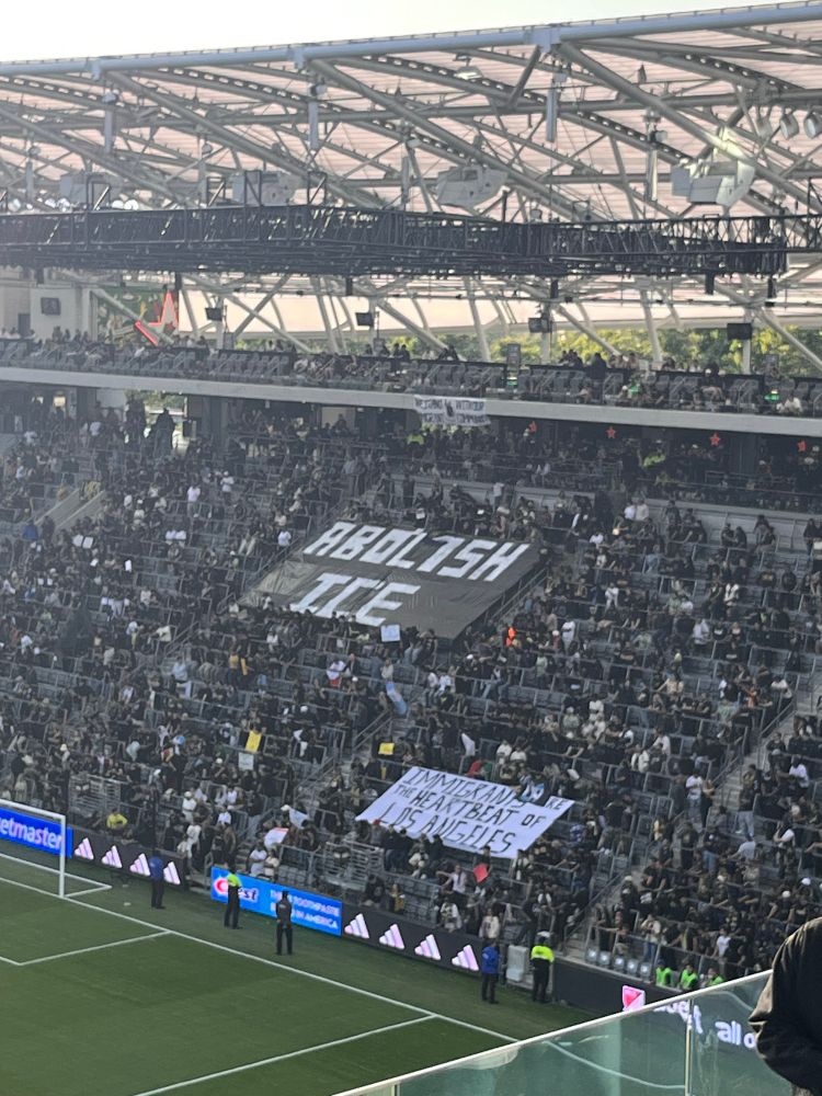Supporters section at an LAFC game holding a sign that says “Abolish Ice” and “immigrants are the heartbeat of LA”