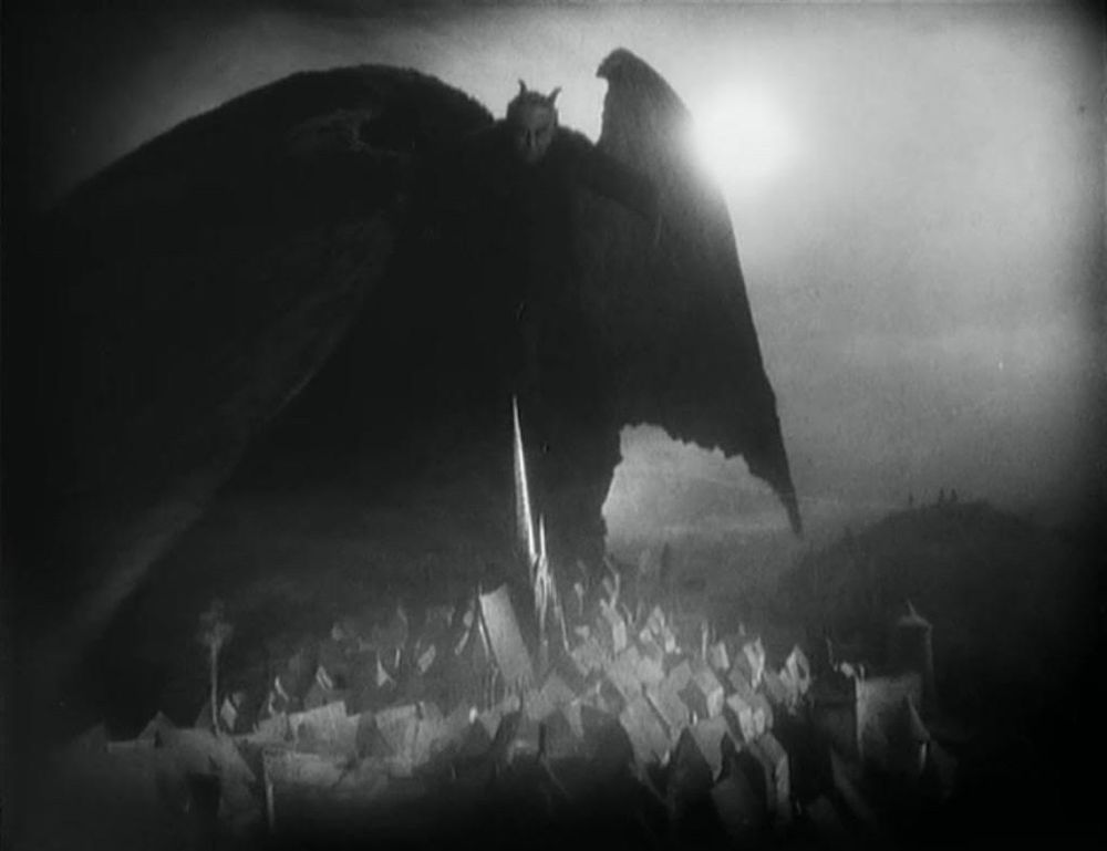 A horned and winged demon stands over Faust' mediaeval home town, misty moonlight glints behind one vast wing. Still from the 1926 movie. 
