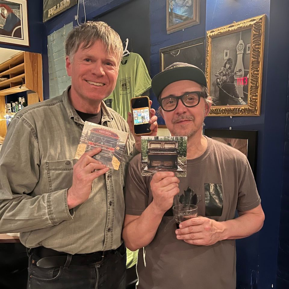 Danny Michel and I with the two CDs that were missing from my collection. The photo is two men facing the camera, each holding a CD, in front of a merch table after a concert at the Gordon Best Theatre. 