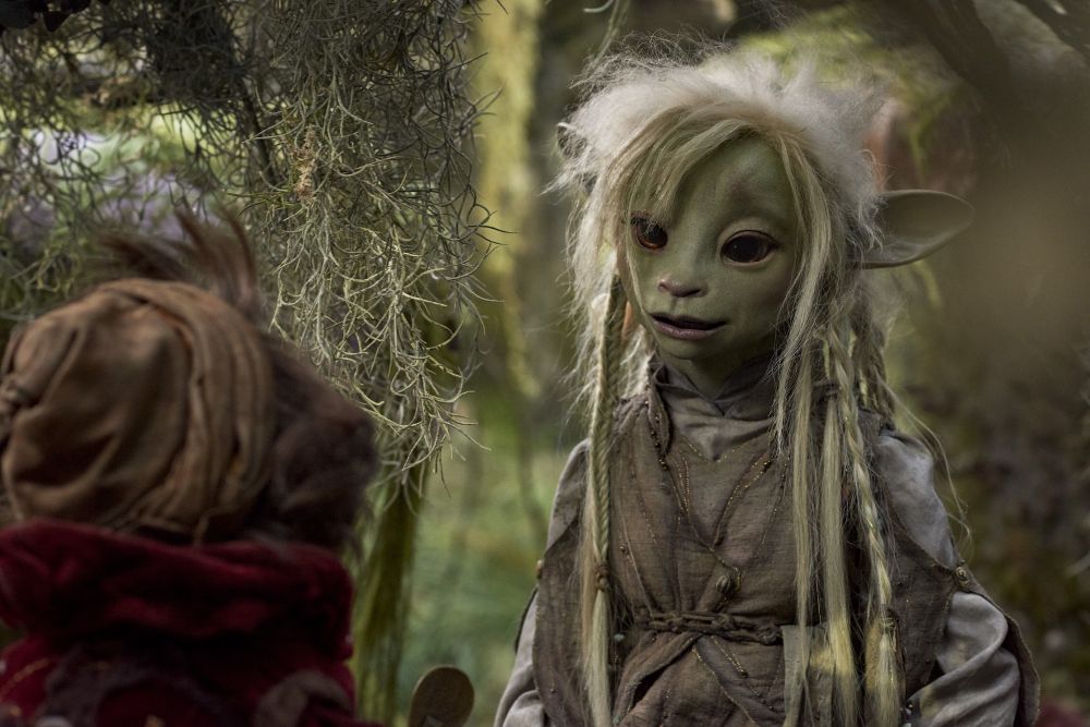 A screen cap from The Dark Crystal: Age of Resistance featuring one of the Gelfling puppets, a Grottan named Deet. She has blonde hair, large dark eyes, and faintly green-tinted skin from living underground.