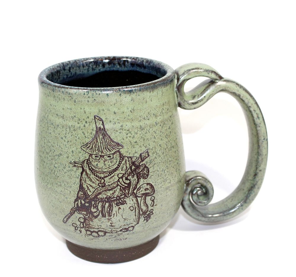 A mossy green mug with an ornate spiral handle and an anthropomorphic frog wizard wearing a robe and leaving toadstool where it walks engraved on the mug