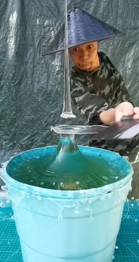 Freeze frame of a man in a black coolie hat slicing through the splash from a bucket of water. Its very possible that he has a body, but one cannot tell because all that is visible are his head and a hand holding the cleaver thats being used to slice through the water