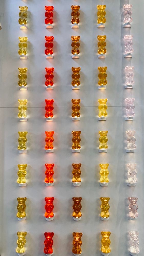 An einer weißen Wand sind eine größere Anzahl von Gummibärleuchten, nach Farben sortiert, angebracht. Es handelt sich um fünf Spalten und acht Reihen. Die Farben sind in Spalten sortiert: gelb, rot, orange, gelb, weiß.