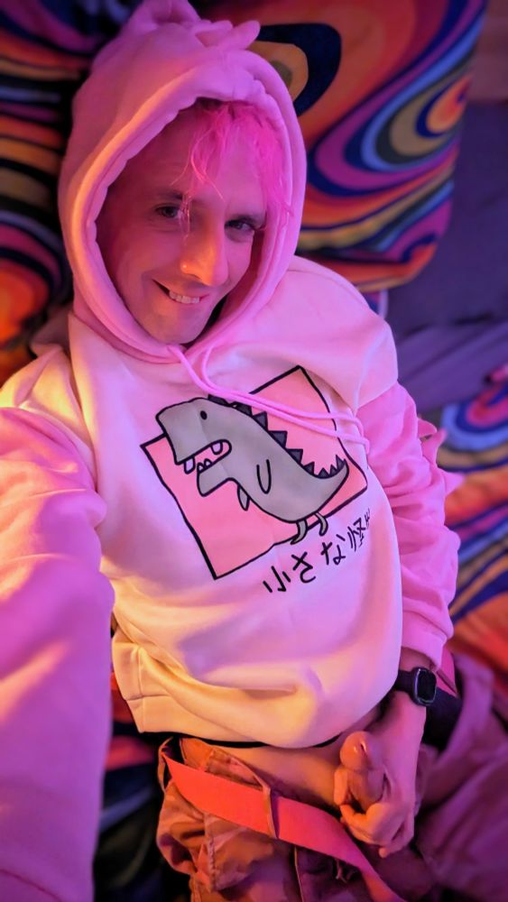 Kittz (a non-binary person with pink hair) laid on their bed, smiling mischeviously in a pink & white dinosaur hoodie, their pink camoflauge combat trousers tugged open, showing off their dick for the camera.