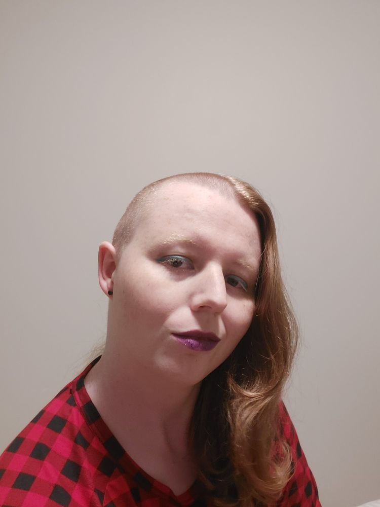 A ginger fem-leaning individual with a side shave is in front of a white wall, with teal eyeliner and deep purple lipstick, showing a half-smile. They are wearing a red and black checkered top, and black earrings, and the picture cuts off at one shoulder and the middle of the other arm.