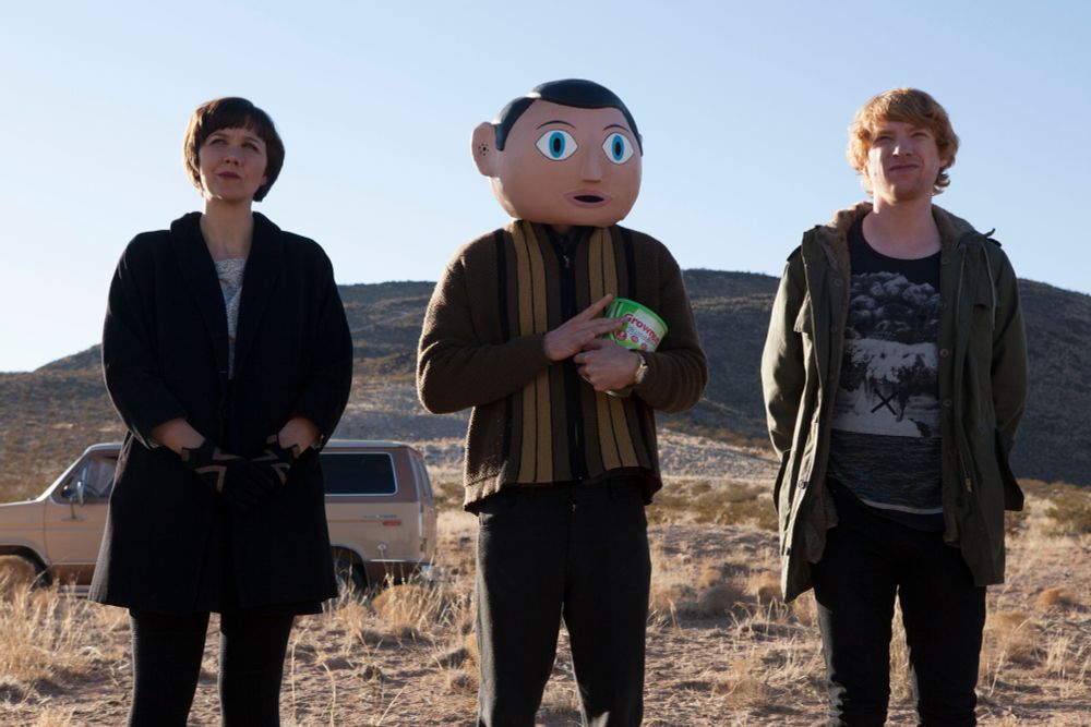 Scene from the film Frank (2014). Three people standing in a desert, one wearing a large papier-mâché mask over his head.