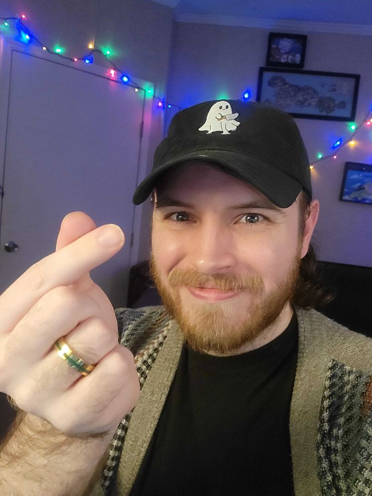 Picture of myself wearing a black hat with a ghost on it. Wearing a black t shirt with a tan cardigan. Holding up a lil "love" sign with my thumb across my index finger.