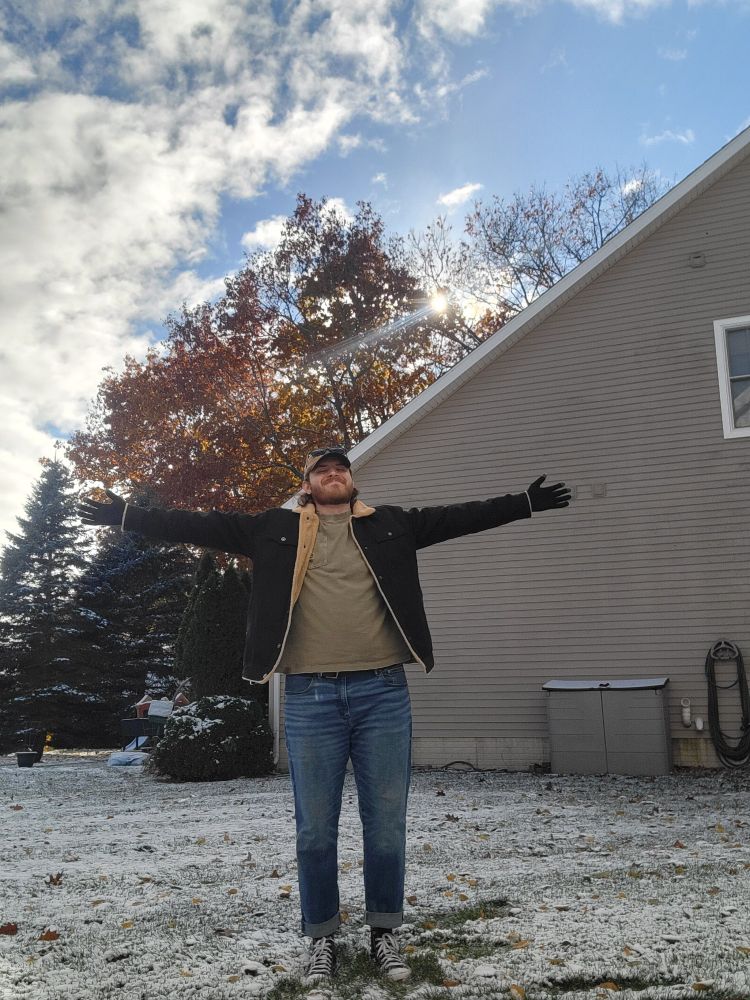 Picture of me with arms spead out wide smiling among some snowy grass. I'm wearing a green tshirt with a black jacket. Blue jeans and black converse. 