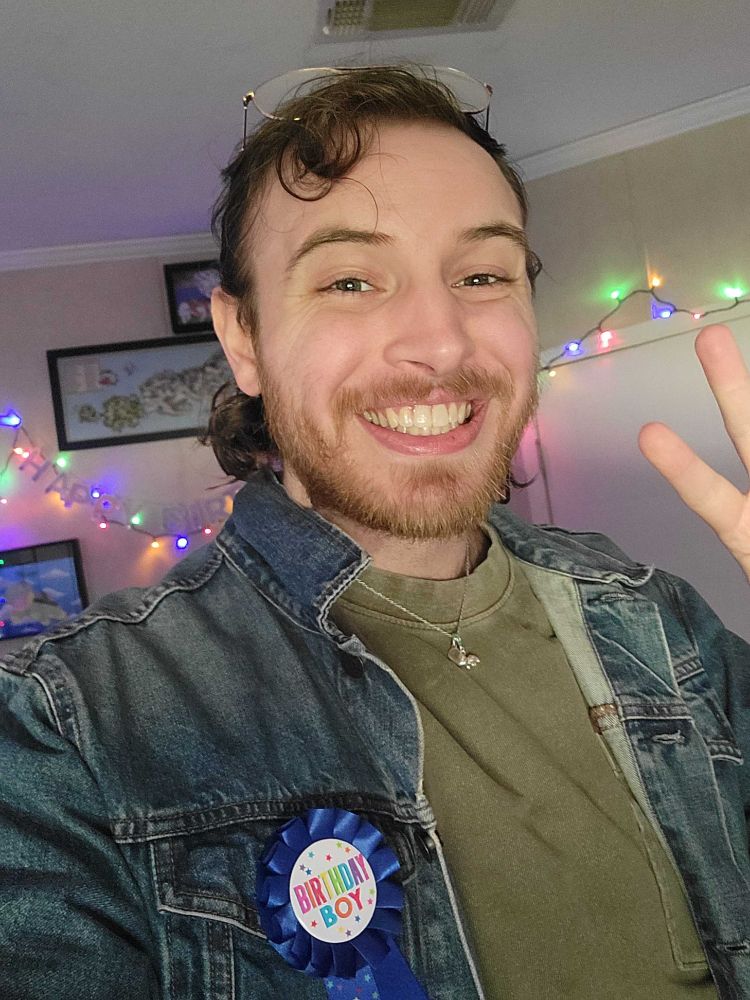 A selfie of Twig! He has a denim blue jacket with a green shirt. The jacket has a blue badge that says "Birthday boy" on it! He's holding up a peace sign as well!!