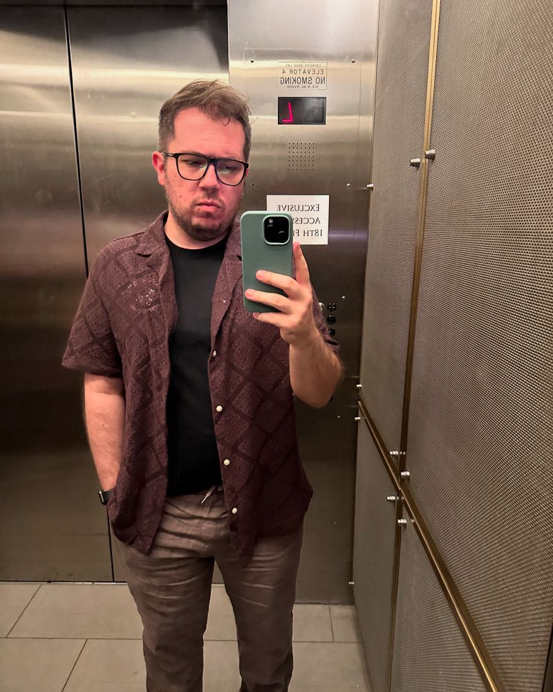 Selfie standing in a lift wearing a black tshirt, purple knitted overshit and linen trousers