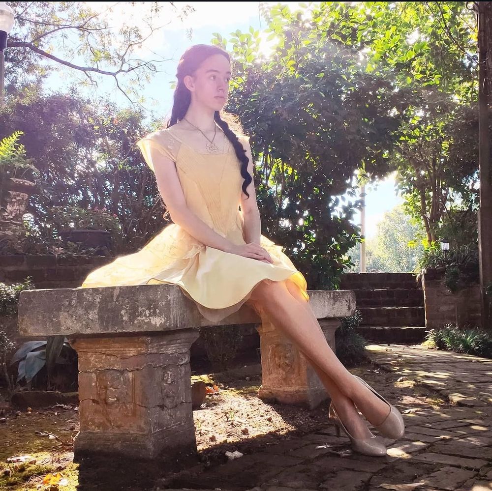 Cosplayer dressed as Belle from Beauty and the Beast, in a knee length dress, sitting on a bench.