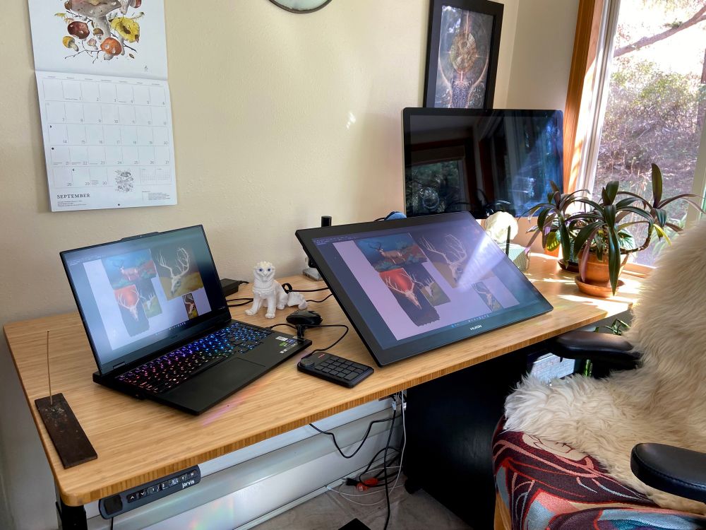 A bamboo desk with a laptop, drawing tablet, monitor, and other items strewn across it in sunlight by a window with houseplants. A chair with a sheepskin and blanket and mess of wires can also be seen.