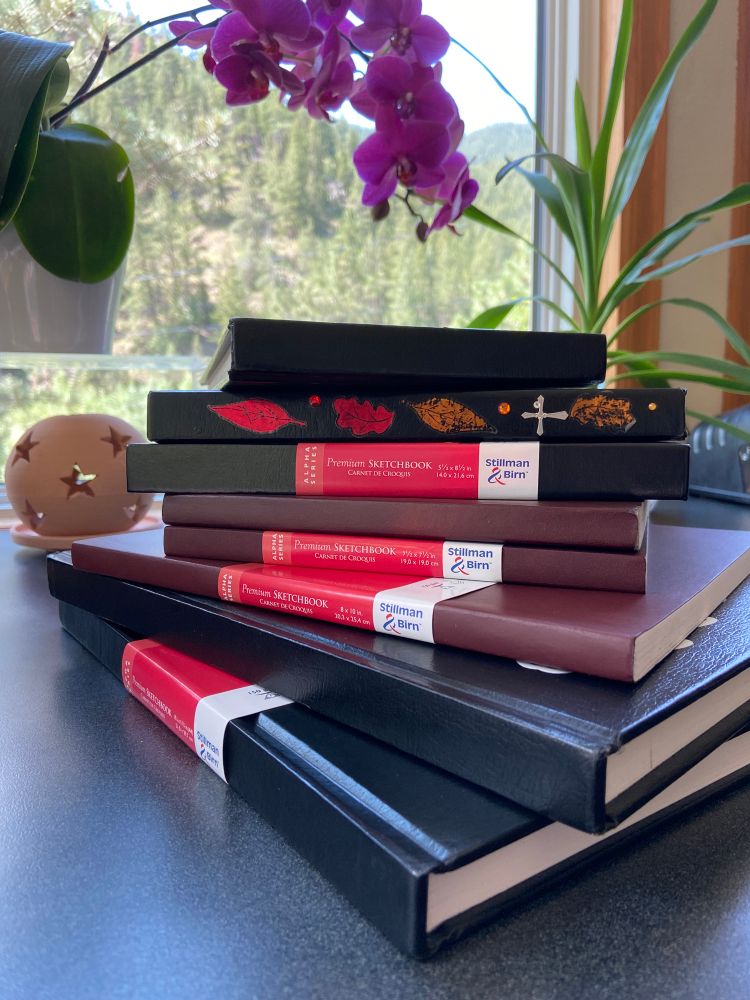 A pile of sketchbooks in different sizes and bindings on a desk.