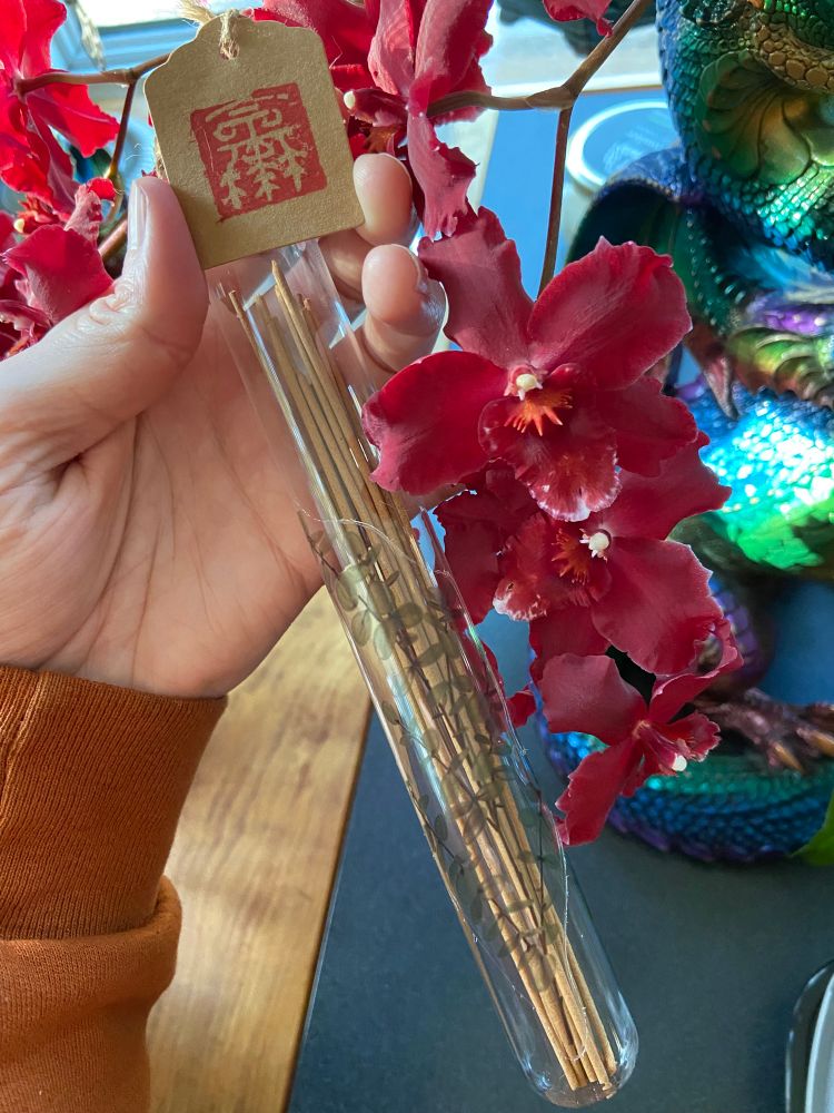 A hand holding a glass tube with incense sticks inside and with a red stamped label. Red Oncidium orchid flowers nestle against the tube. A desk is in the background. 