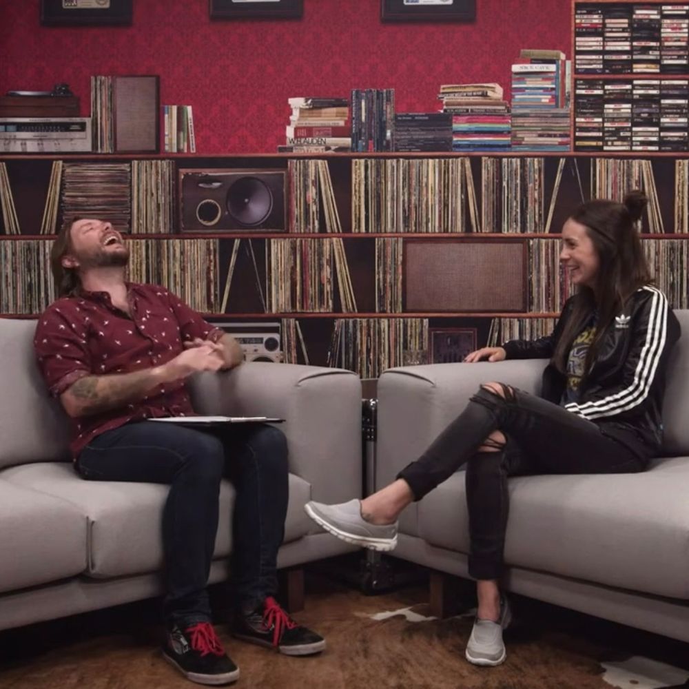 Two people sit on a grey couch in front of shelves full of records and CDs. The people are sitting facing each other, one has his head thrown back in laughter, the other, Amy Shark, is laughing much more demurely. 