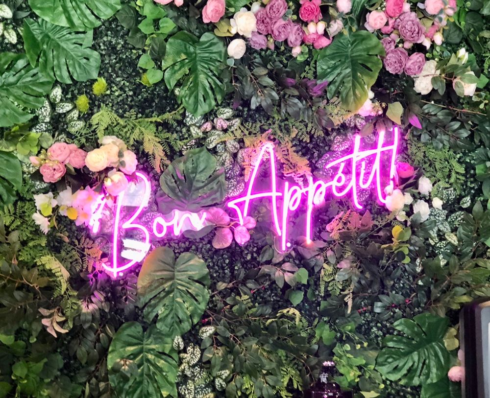 Pink neon letters on a green floral background reading “Bon Appetit!”, a cute photo spot at a vegan patisserie ID-Eclair