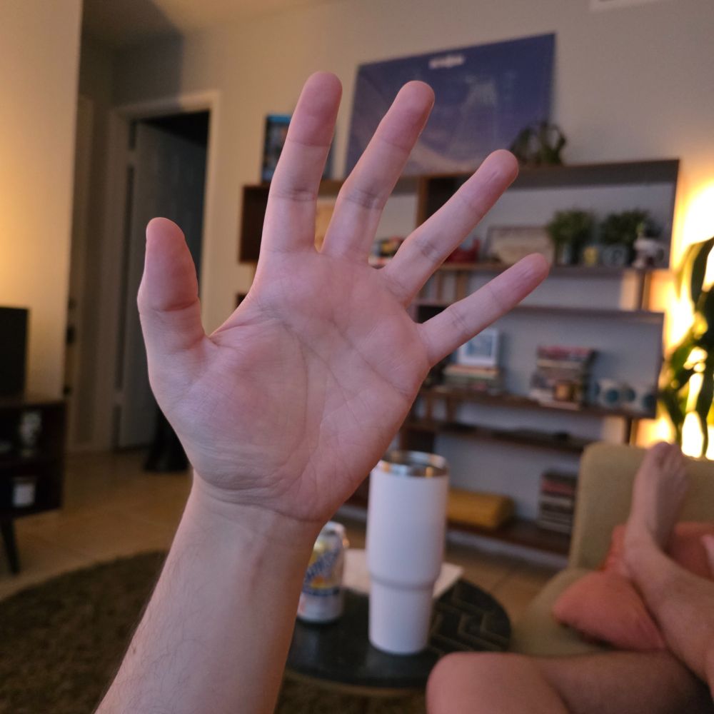 A person's left hand, palm open, is shown in the foreground with a living room visible in the background.