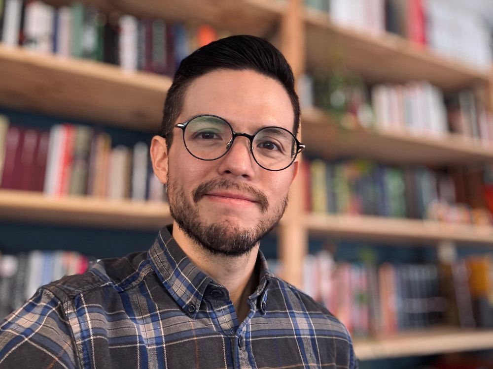 Selfie in front of my bookshelf, wearing glasses and a blue plaid shirt. 