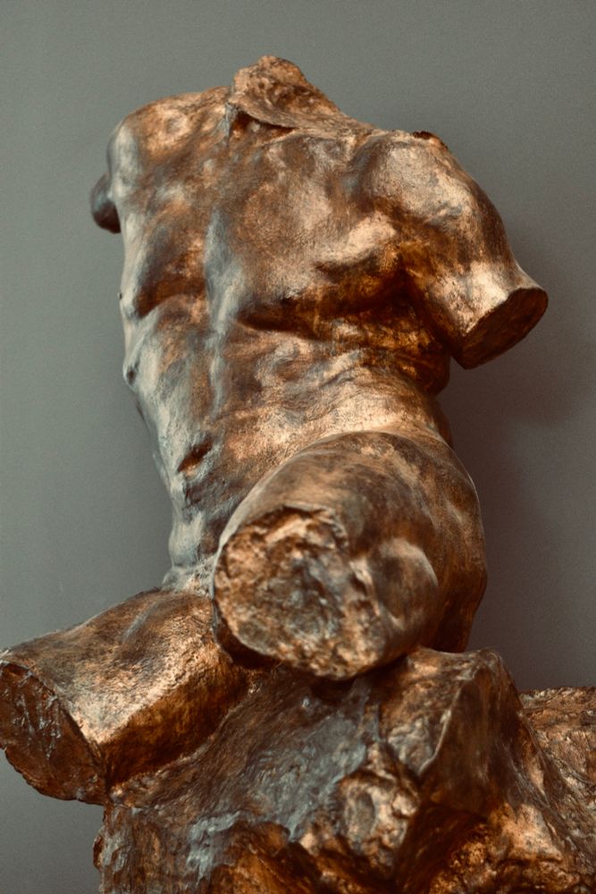 A sculpture of a nude male torso. It is missing its limbs and head. It is made of a copper like material. Sturdy. Strong. Beautiful. 