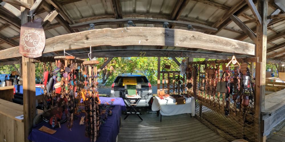 A panoramic photo of a leather shop in a wooden booth. Bags hang on racks. Hair pins hang on hooks. Belts and things to hang on belts are on display. Little leather critters are also on display.